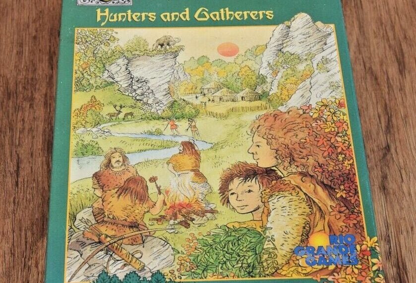 A board game box for "Carcassonne: Hunters and Gatherers" on a wooden surface, featuring illustrated prehistoric people and a lush landscape.