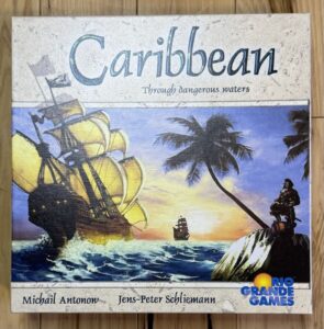 Box cover of the "Caribbean" board game featuring a large sailing ship on turbulent seas with smaller ships in the background, a tropical setting with palm trees, and a pirate figure on a rock in the foreground. The text on the cover includes the game's title and subtitle 'Through dangerous waters' and the names 'Michail Antonow' and 'Jens-Peter Schliemann' with the 'Rio Grande Games' logo at the bottom right.