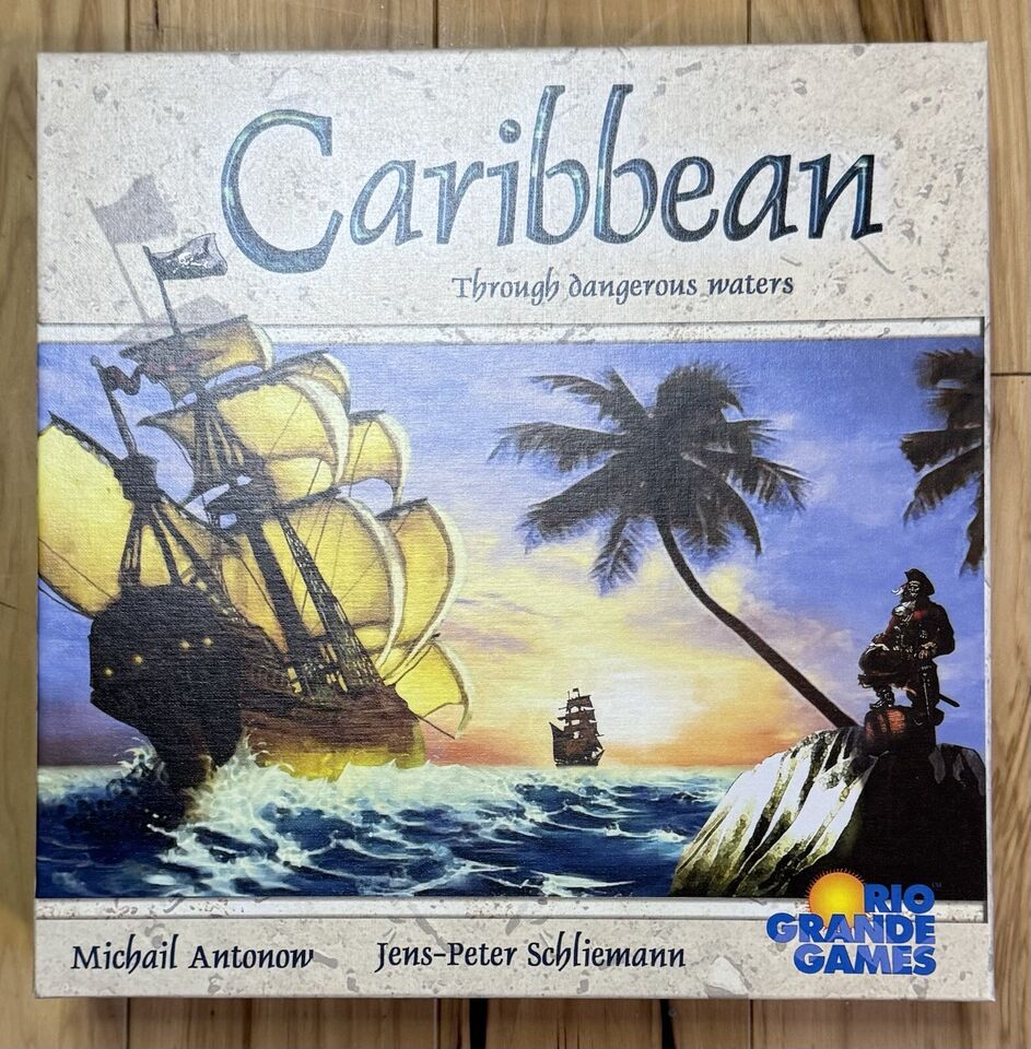 Box cover of the "Caribbean" board game featuring a large sailing ship on turbulent seas with smaller ships in the background, a tropical setting with palm trees, and a pirate figure on a rock in the foreground. The text on the cover includes the game's title and subtitle 'Through dangerous waters' and the names 'Michail Antonow' and 'Jens-Peter Schliemann' with the 'Rio Grande Games' logo at the bottom right.