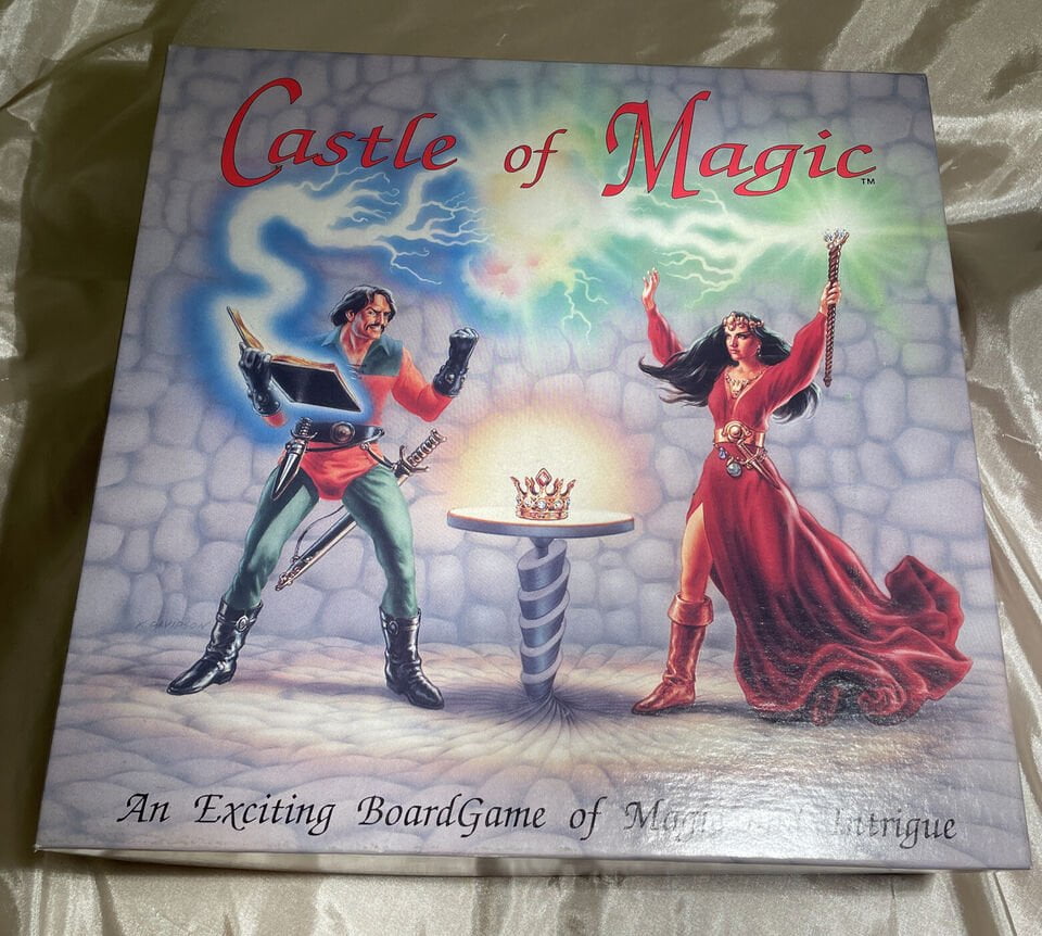 A board game titled "Castle of Magic" featuring artwork with two fantasy characters — one reading from a magical book with energy emanating from it, and the other, a female figure, holding a staff with a swirling magical aura above. A crown on a pedestal appears in the center, all set against a stone floor and a mystical blue and green cloudy background. The subtitle reads, "An Exciting Board Game of Magic & Intrigue."
