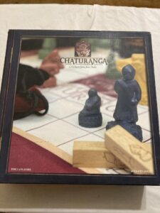 The image displays a slightly faded box cover of a board game named "CHATURANGA, A Classic Game from India," recommended for 2-4 players, ages 8 to adult. The cover features a close-up view of two carved game pieces on a chess-like board, with a blurred background suggesting a person’s hands in motion.