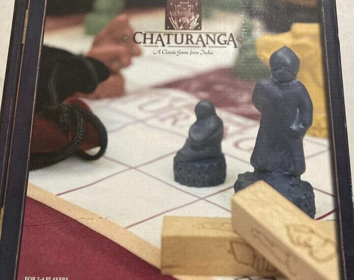 The image displays a slightly faded box cover of a board game named "CHATURANGA, A Classic Game from India," recommended for 2-4 players, ages 8 to adult. The cover features a close-up view of two carved game pieces on a chess-like board, with a blurred background suggesting a person’s hands in motion.