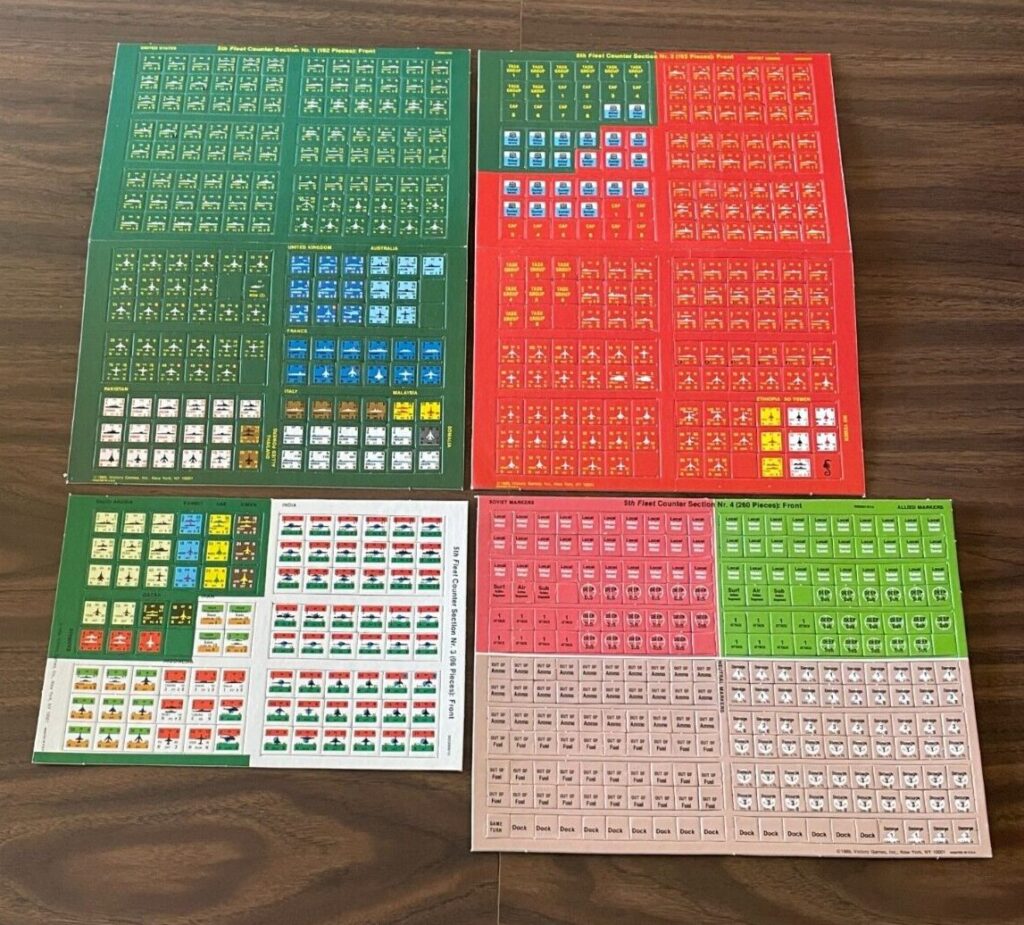 Four sheets of colorful board game counters laid out on a wooden floor, with each sheet featuring a different color (green, red, white, and brown) and various symbols and numbers on each counter.