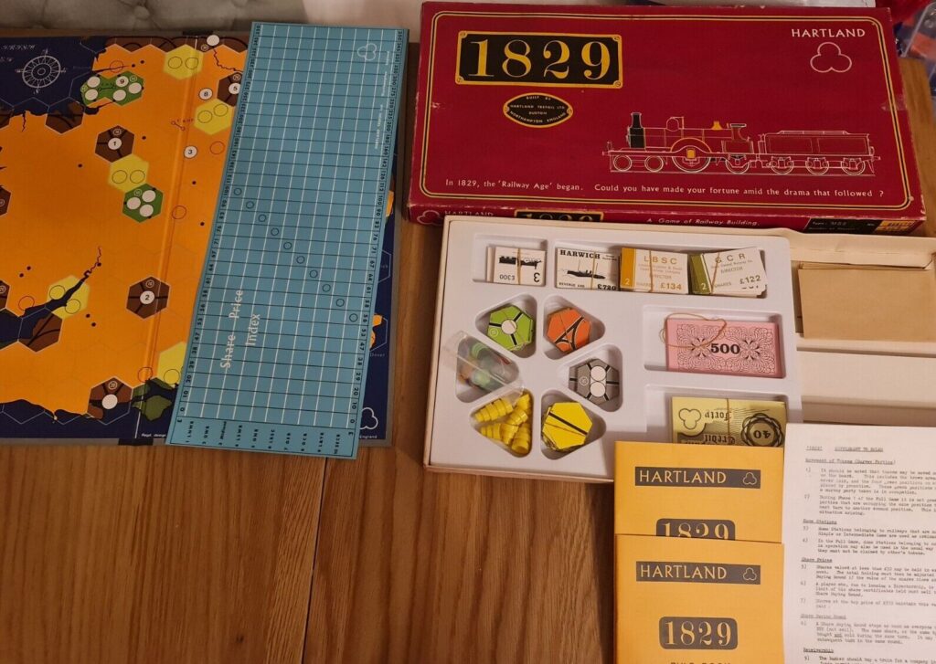 A tabletop setup displaying components of the board game 1829 including a map board, stock price index, various game cards, currency notes, game pieces, the game box with a vintage locomotive illustration, and instructions laid out on a wooden surface.