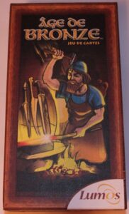Box for "Age de Bronze" card game featuring an illustration of a bearded man in ancient attire forging a weapon by a fire.